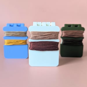 May include: Three thread organizers in blue, white, and green, each holding different colored embroidery floss. The floss colors include brown, yellow, and burgundy. The organizers are on a pink background.
