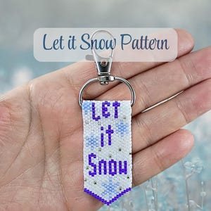May include: A rectangular beaded keyring featuring a white background with the text Let it Snow in purple letters. The design includes light blue snowflake accents and silver metallic beads. It is attached to a silver metal lobster clasp.