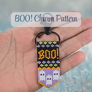 May include: A rectangular woven keychain featuring the text BOO! in orange on a black background, accented with green and purple geometric patterns and white ghost figures at the bottom, attached to a black metal lobster clasp and ring hardware.