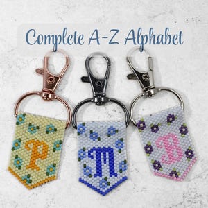 May include: Three beaded keychains featuring the letters P, M, and B in floral patterns with rose gold, gunmetal, and silver lobster clasps. The text Complete A-Z Alphabet is visible at the top. Each pendant has a pointed bottom edge.