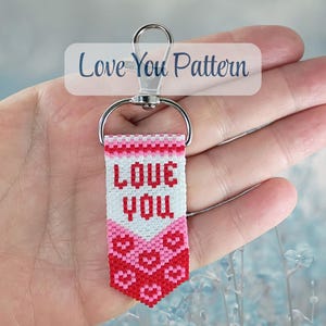 May include: A rectangular beaded keyring featuring the text LOVE YOU in red on a white background, with a pink and red heart pattern below. The keyring is attached to a silver-coloured metal lobster clasp and a semi-circular metal connector.