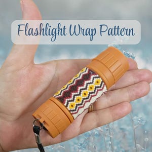 Puede incluir: Una pequeña linterna naranja con una envoltura tejida de patrón geométrico en colores amarillo, marrón oscuro y crema. El texto Flashlight Wrap Pattern aparece sobre el objeto, que incluye una correa de muñeca negra en la base.