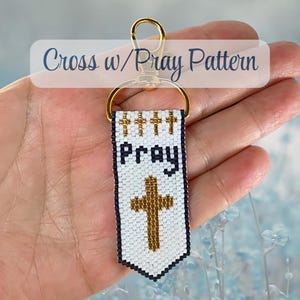 Puede incluir: Un llavero rectangular de cuentas con fondo blanco y borde azul oscuro. El diseño incluye cuatro pequeñas cruces doradas en la parte superior, la palabra "Pray" en azul oscuro y una cruz dorada más grande debajo. Tiene un cierre de metal dorado.