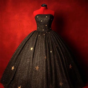May include: A strapless black gown with gold star embellishments. The dress has a full skirt and is displayed on a red dress form. The fabric has a subtle shimmer, and the stars are scattered across the bodice and skirt.