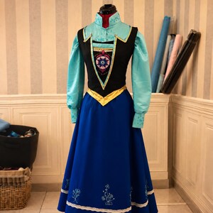 May include: A long-sleeved dress featuring a light blue high-neck bodice with floral embroidery, a black vest-style overlay with a central geometric emblem, a gold waistband, and a floor-length royal blue skirt with white lace and floral embroidery.