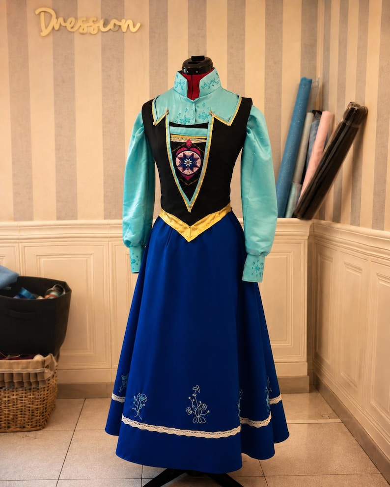 May include: A long-sleeved dress featuring a light blue high-neck bodice with floral embroidery, a black vest-style overlay with a central geometric emblem, a gold waistband, and a floor-length royal blue skirt with white lace and floral embroidery.