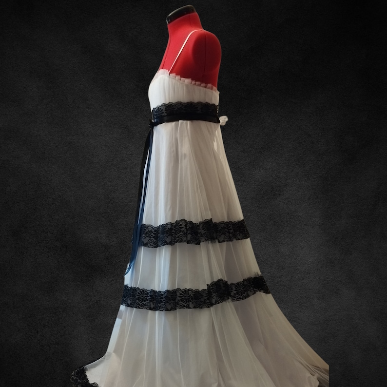 Puede incluir: Un vestido de tul blanco con ribetes de encaje negro y una cinta de raso negra. El vestido tiene un corpi&ntilde;o ajustado y una falda fluida.