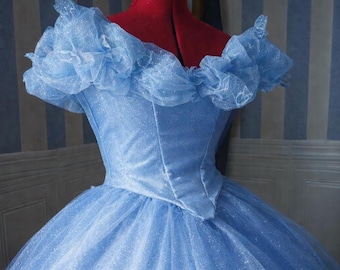May include: A light blue, glittery ball gown with off-the-shoulder ruffled sleeves. The bodice is fitted, and the skirt is full and layered. The dress is displayed on a red dress form against a striped background.
