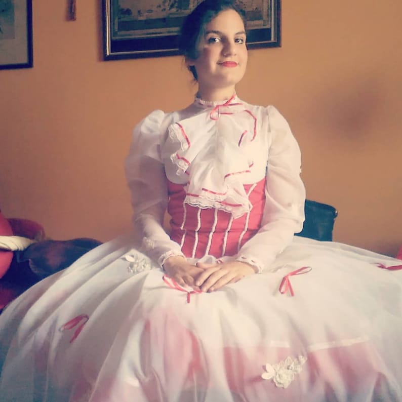 May include: A woman wearing a white dress with red trim and a white blouse with a red bow. The dress has a full skirt and the woman is sitting on a chair.