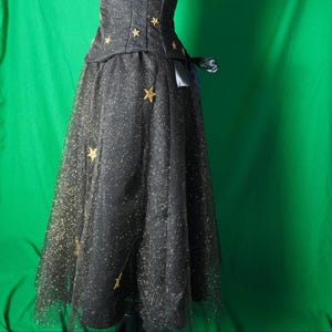 May include: A black dress with a fitted bodice and a flowing skirt. The dress is embellished with gold star appliqu&eacute;s and a layer of glittery fabric. A bow detail accents the waist.
