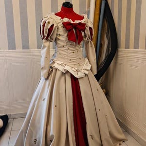 May include: A cream-colored ball gown with red accents and a large red bow. The dress features long sleeves with red puffed shoulders, a fitted bodice, and a full skirt. Gold floral patterns are scattered across the fabric.