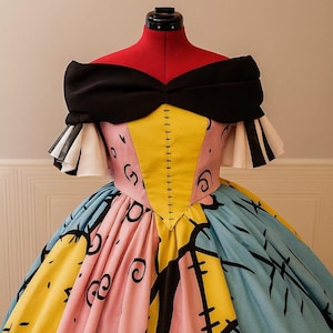 Könnte beinhalten: Ein farbenfrohes schulterfreies Kleid auf einer roten Schneiderpuppe. Das Oberteil ist ein Patchwork aus rosa, gelbem und schwarzem Stoff mit einer schwarzen Schleife am Ausschnitt. Der Rock ist eine Mischung aus blau, gelb und rosa mit schwarzen grafischen Designs.