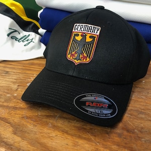Op de afbeelding: Zwarte baseballpet met een zwart en geel geborduurd Duits wapenschild met de tekst "Germany". De pet heeft een zwart Flexfit-label met de tekst "The Original Flexfit L/XL It Fits Your Life."