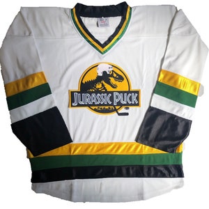 May include: White hockey jersey with black, green, and gold stripes. The jersey features a large logo of a dinosaur skeleton with a hockey helmet on its head. The logo is on a yellow circle with the text "Jurassic Puck" below it.