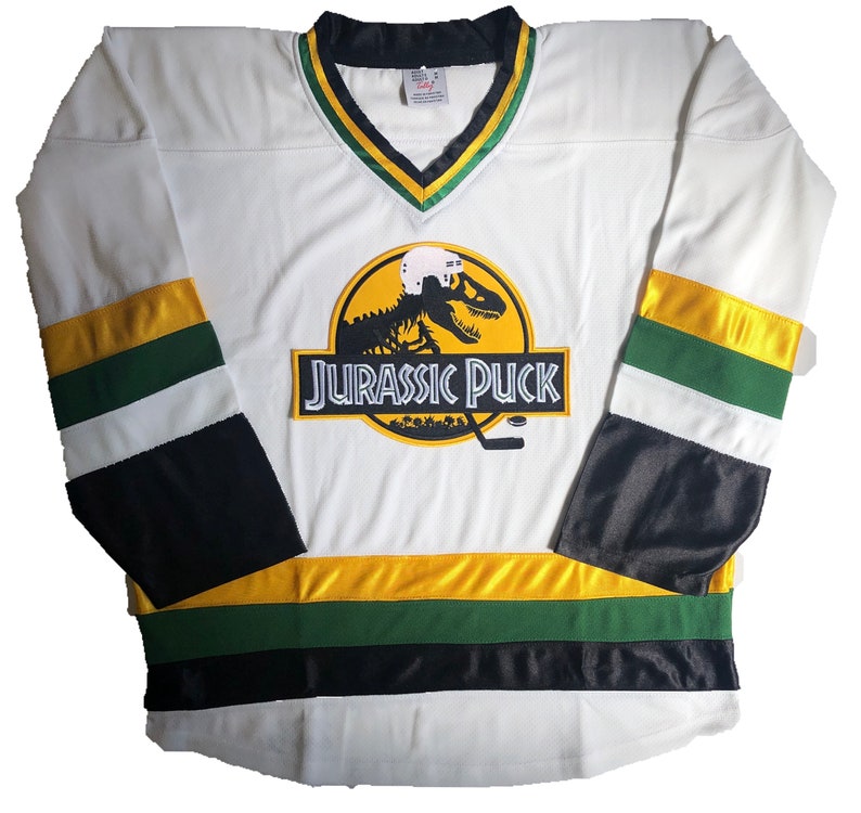 May include: White hockey jersey with black, green, and gold stripes. The jersey features a large logo of a dinosaur skeleton with a hockey helmet on its head. The logo is on a yellow circle with the text "Jurassic Puck" below it.