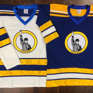 Op de afbeelding: Twee hockeytruien, een witte met blauwe en gouden strepen en een blauwe met witte en gouden strepen. Beide truien hebben een logo van een scheidsrechter met het woord "OFFSIDERS" eronder.