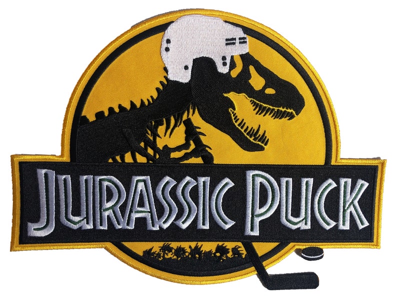 May include: A black and yellow embroidered patch with a dinosaur skeleton wearing a hockey helmet. The text "Jurassic Puck" is below the dinosaur. A hockey stick and puck are at the bottom of the patch.