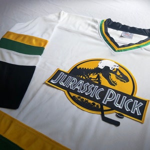 May include: White hockey jersey with green and gold stripes. The front of the jersey features a large patch with a dinosaur skeleton wearing a hockey helmet and the text "Jurassic Puck".
