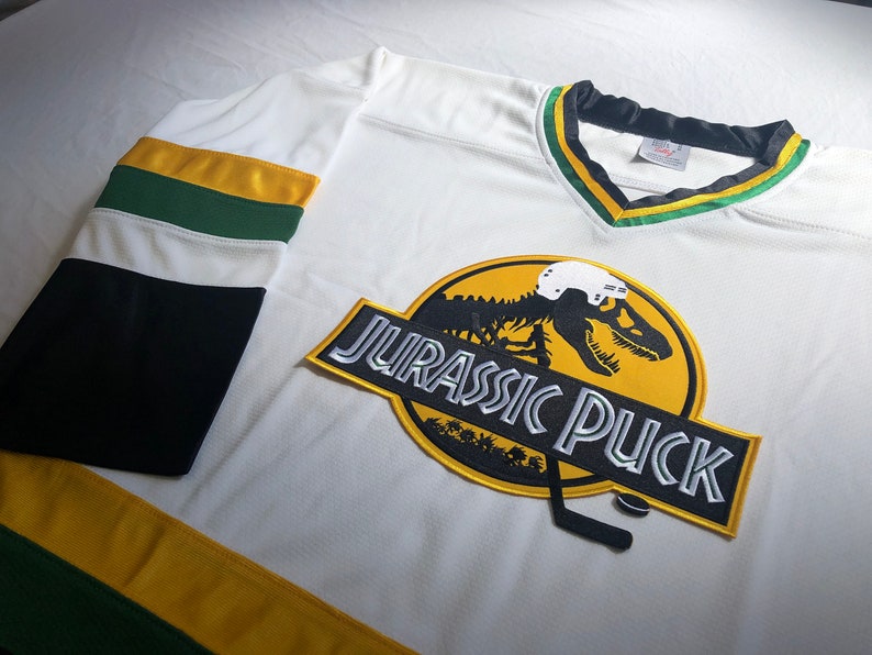 May include: White hockey jersey with green and gold stripes. The front of the jersey features a large patch with a dinosaur skeleton wearing a hockey helmet and the text "Jurassic Puck".