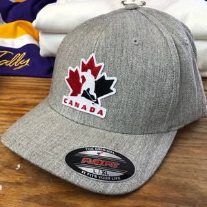 May include: Gray baseball cap with a red, white, and black Canadian maple leaf logo featuring a hockey player. The logo says "CANADA". The cap has a Flexfit label that says "The Original Flexfit L/XL It Fits Your Life."