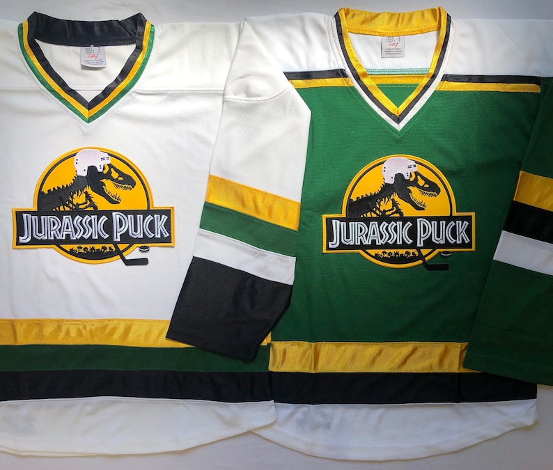 May include: Two hockey jerseys with a dinosaur skeleton graphic and the text "Jurassic Puck". One jersey is white with green and gold stripes, the other is green with white and gold stripes.