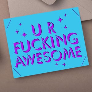 May include: A square greeting card with the words "UR FUCKING AWESOME" in purple, stylized font on a light blue background. The card has star accents and is on a brown surface.