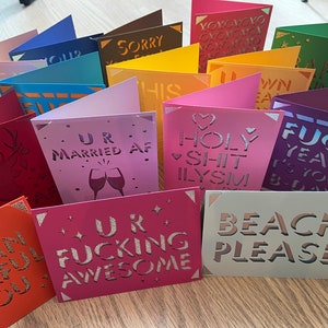 May include: A collection of colorful greeting cards with various humorous and heartfelt messages. The cards feature laser-cut designs and glitter accents. Some of the messages include "UR MARRIED AF", "HOLY SHIT ILYSM", "FUCK YEAH IT'S YOUR BIRTHDAY", and "UR FUCKING AWESOME".