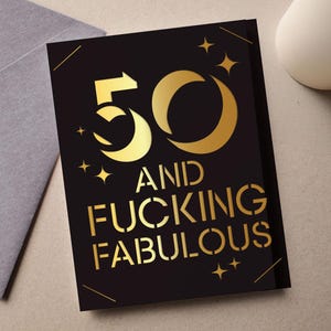 Könnte beinhalten: Eine schwarze Grußkarte mit goldfarbenen Buchstaben und Akzenten. Auf der Karte steht "50 AND FUCKING FABULOUS" in einer Schablonenschrift. Die Zahl 50 ist mit einem Mond und Pfeildesign stilisiert. Die Karte liegt neben einem grauen Umschlag.