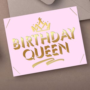 May include: A pink card with gold lettering that reads "BIRTHDAY QUEEN" and a gold crown illustration. The card is on a brown surface with a gift box and envelope in the background.