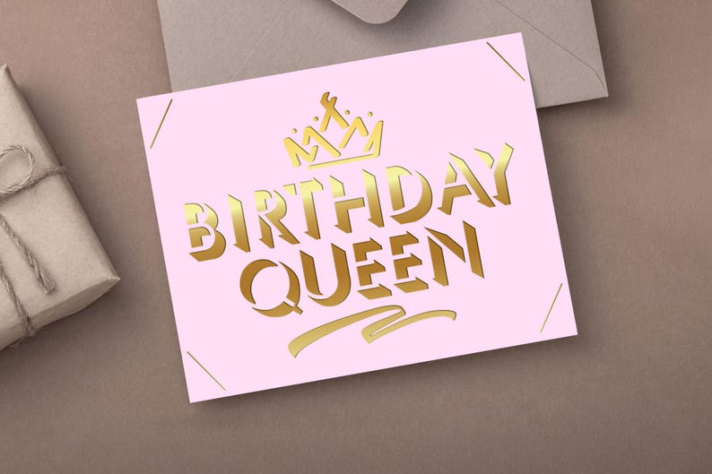 May include: A pink card with gold lettering that reads "BIRTHDAY QUEEN" and a gold crown illustration. The card is on a brown surface with a gift box and envelope in the background.