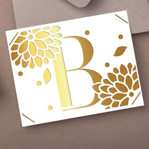May include: A white card with a gold letter "B" and floral cutouts. The design includes gold accents like dots and leaf shapes. The card is on a brown surface, with a wrapped gift and envelope in the background.