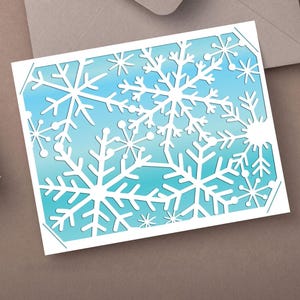 May include: A white card with a laser-cut snowflake design. The card has a blue and white gradient background. The snowflakes are of varying sizes and shapes. The card is on a brown surface with a gift and envelope.