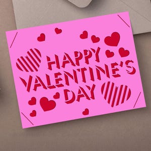 May include: A pink Valentine's Day card with the words "HAPPY VALENTINE'S DAY" cut out in red. The card features red hearts and striped heart designs. The card is on a neutral surface.