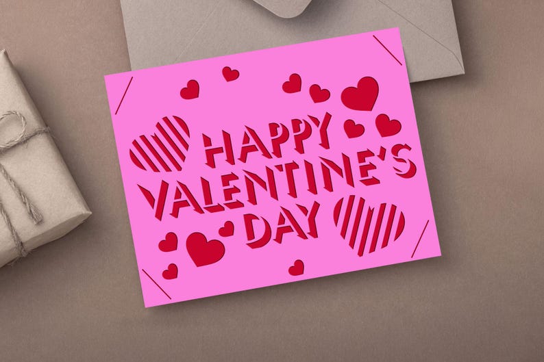 May include: A pink Valentine's Day card with the words "HAPPY VALENTINE'S DAY" cut out in red. The card features red hearts and striped heart designs. The card is on a neutral surface.