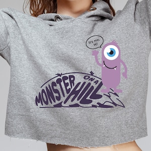 Pode incluir: Uma camisa de moletom cinza com capuz com uma estampa gráfica de um monstro roxo com um olho e o texto "Monster on a Hill". O monstro está dizendo "Sou eu, Oi!" em um balão de fala.