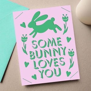 May include: A pink greeting card with the text "SOME BUNNY LOVES YOU" in green, along with a green bunny silhouette, hearts, and floral accents. A teal envelope is visible in the background. The card is likely for Easter or a similar occasion.