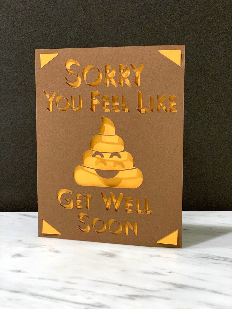May include: A brown get-well card with the words "Sorry You Feel Like" and "Get Well Soon" cut out. A smiling poop emoji is in the center. The card has gold accents in the corners.