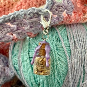 May include: A small glass bottle charm filled with dried herbs and gold flakes, topped with a cork and purple wax. The charm is attached to a silver clasp, hanging against a backdrop of pastel-colored yarn and crochet.