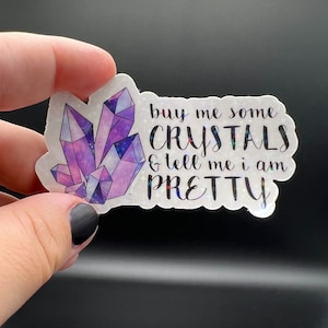 May include: A holographic sticker with a cluster of purple crystals and the text "buy me some CRYSTALS & tell me i am PRETTY". The sticker has a shiny, iridescent finish and is held by a hand.