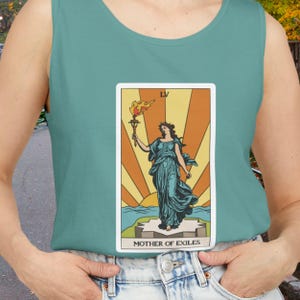 Puede incluir: Camiseta sin mangas turquesa con un diseño de carta del tarot. La carta presenta a una mujer con un vestido turquesa que sostiene una antorcha, con el texto "MOTHER OF EXILES" en la parte inferior. El fondo tiene un diseño de rayos de sol.