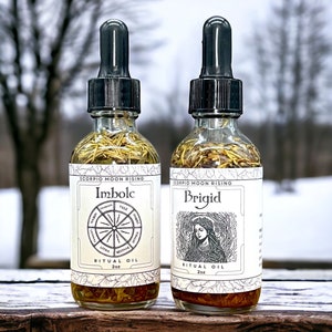 Puede incluir: Dos botellas de vidrio transparente con goteros llenos de un líquido amarillo y hierbas. Las botellas tienen etiquetas blancas con texto negro que dice "Scorpio Moon Rising", "Imbolc", "Ritual Oil", "2oz" y "Brigid".