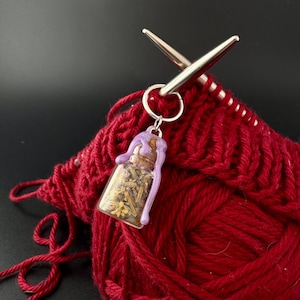 May include: A small glass bottle charm filled with dried herbs and a purple wax seal, attached to a silver keyring. The charm is set against a backdrop of red yarn and knitting needles, suggesting a craft-related theme.