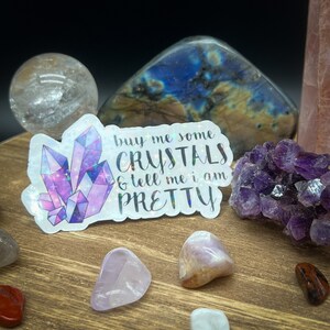 May include: A holographic sticker with the text "buy me some CRYSTALS & tell me i am PRETTY" and a purple crystal design. The sticker is surrounded by various crystals and gemstones, including a clear quartz sphere and amethyst.