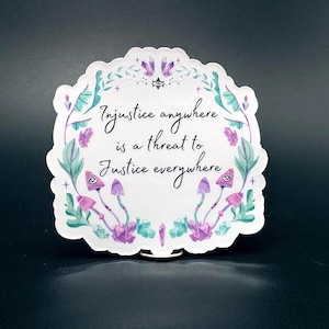 May include: A white sticker with a scalloped edge, featuring the quote "Injustice anywhere is a threat to Justice everywhere." The design includes watercolor illustrations of mushrooms, crystals, and foliage in shades of purple, teal, and green.
