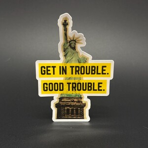 May include: A sticker featuring the Statue of Liberty with the text "GET IN TROUBLE. GOOD TROUBLE." The sticker has a white border and a yellow background for the text. The Statue of Liberty is green and brown.