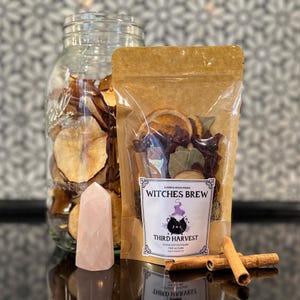May include: A collection of items for a witch-themed product. Includes a clear jar filled with dried apple slices, a package labeled "Witches Brew", a pink crystal, and cinnamon sticks. The package contains dried fruit and herbs.