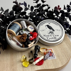 May include: A silver tin of Magick Crow Confetti with a crow illustration on the lid, and various small objects inside, including a spool of blue thread, a thimble, a toy car, and a red heart. A tarot card and other trinkets are also visible.