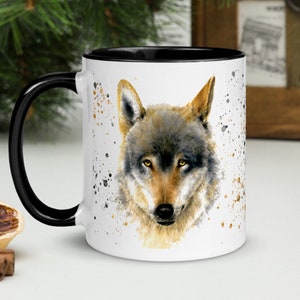 K&ouml;nnte beinhalten: Wei&szlig;e Keramiktasse mit schwarzem Innenraum und Henkel. Die Tasse zeigt ein Aquarellgem&auml;lde eines Wolfsgesichts mit Braun-, Grau- und Goldt&ouml;nen. Das Design umfasst Farbspritzer.