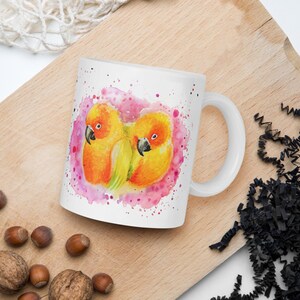 Könnte beinhalten: Weiße Keramiktasse mit einer Aquarell-Illustration von zwei gelben und orangefarbenen Papageien, die sich auf einem rosa Hintergrund mit weißen Spritzern aneinanderkuscheln.