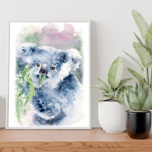 May include: Watercolour painting of a koala bear eating eucalyptus leaves. The artwork features shades of blue and grey, with a touch of pink in the background. The framed print is displayed on a wooden surface, with potted plants in the background.